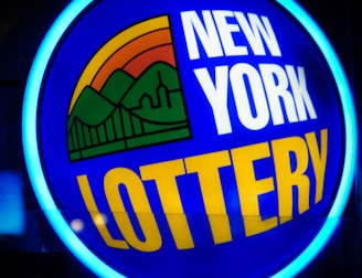 A bright, circular neon sign with a blue background, featuring the words 'New York Lottery' in bold white and yellow letters. The sign includes a colorful illustration of a landscape with green mountains, an orange-red sunset, and a bridge, all encapsulated within the circle.