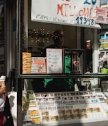 A child holding an ice cream cone stands outside a small kiosk or lottery ticket booth. The booth displays various colorful lottery tickets in the window. There are signs advertising a significant lottery prize amount in Spanish. A person can be seen partially obscured inside the booth, and plants are visible around the area, suggesting an outdoor setting.