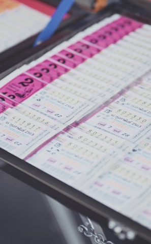 A collection of lottery tickets is organized neatly in a metal tray. The tickets display various numbers and have color-coded sections, primarily in pink and white. Some tickets have vibrant green sections, adding contrast to the overall layout.