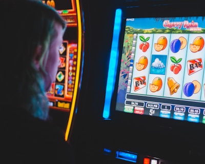 A person is closely observing a digital slot machine with colorful fruit symbols, bars, and a bell. The slot machine screen displays symbols like oranges, plums, cherries, and clouds. The background is dimly lit, creating a focused and somewhat intense atmosphere.