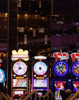 A lively casino scene featuring a row of bright, colorful slot machines with themes like Monopoly, Michael Jackson, and Lord of the Rings. The gaming machines display a variety of flashing lights and digital screens, creating an energetic and exciting atmosphere. Reflections of the lights are visible in glossy surfaces above the machines.