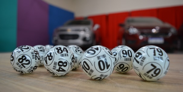 A collection of white lottery balls with black numerals scattered on a wooden surface. Two vehicles are blurred in the background, parked in a room with multicolored walls including teal, purple, blue, and red panels.