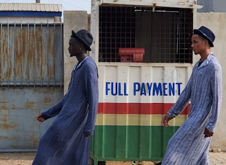 Two individuals are walking past a small kiosk with signage for the National Lottery Authority. The kiosk, decorated with a sign that reads 'Full Payment,' features yellow, green, red, and white colors. The individuals are dressed in matching long blue robes and hats, with one wearing black boots and the other white boots.