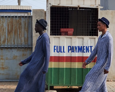 Two individuals are walking past a small kiosk with signage for the National Lottery Authority. The kiosk, decorated with a sign that reads 'Full Payment,' features yellow, green, red, and white colors. The individuals are dressed in matching long blue robes and hats, with one wearing black boots and the other white boots.