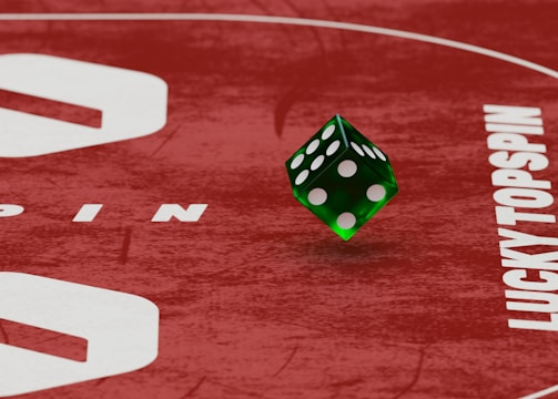 Two green dice with rounded edges are positioned on a red surface that seems to be part of a game board. The surface has white text and numbers on it, including the word 'LUCKYTOPSPIN' in capital letters.