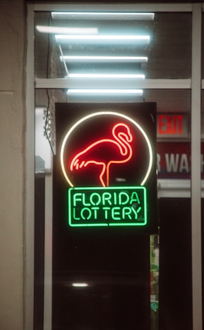 A neon sign depicting a flamingo above the words 'Florida Lottery' is prominently displayed. The sign is illuminated in bright red and green colors, and it is framed by glass panels. An 'EXIT' sign is visible in the background.