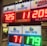 A signboard inside a convenience store displays the current jackpot amounts for various lottery games. There are bright, illuminated numbers in red and green showing the prize amounts: Powerball is at 725 million, Super Lotto Plus at 11 million, Mega Millions at 205 million, Fantasy 5 at 71 thousand, and Daily Derby at 179. In the background, shelves with various drinks and products are visible, along with promotional posters.