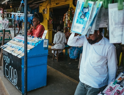 A street vendor stall displaying an array of lottery tickets clipped to a bright blue kiosk. A man in a red shirt leans on the counter, appearing thoughtful, while another man in a white shirt adjusts his hat and walks by. A variety of products hangs from strings above, and a colorful display fills the background with vibrant signs and packets. The scene suggests a busy, lively marketplace.